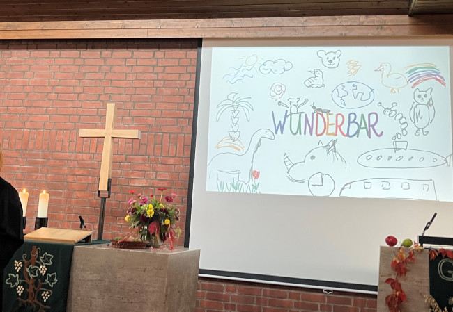 Foto des Altars und einer Projektion mit Beamer: Schriftzug WUNDERBAR und gemalte Tiere drumherum